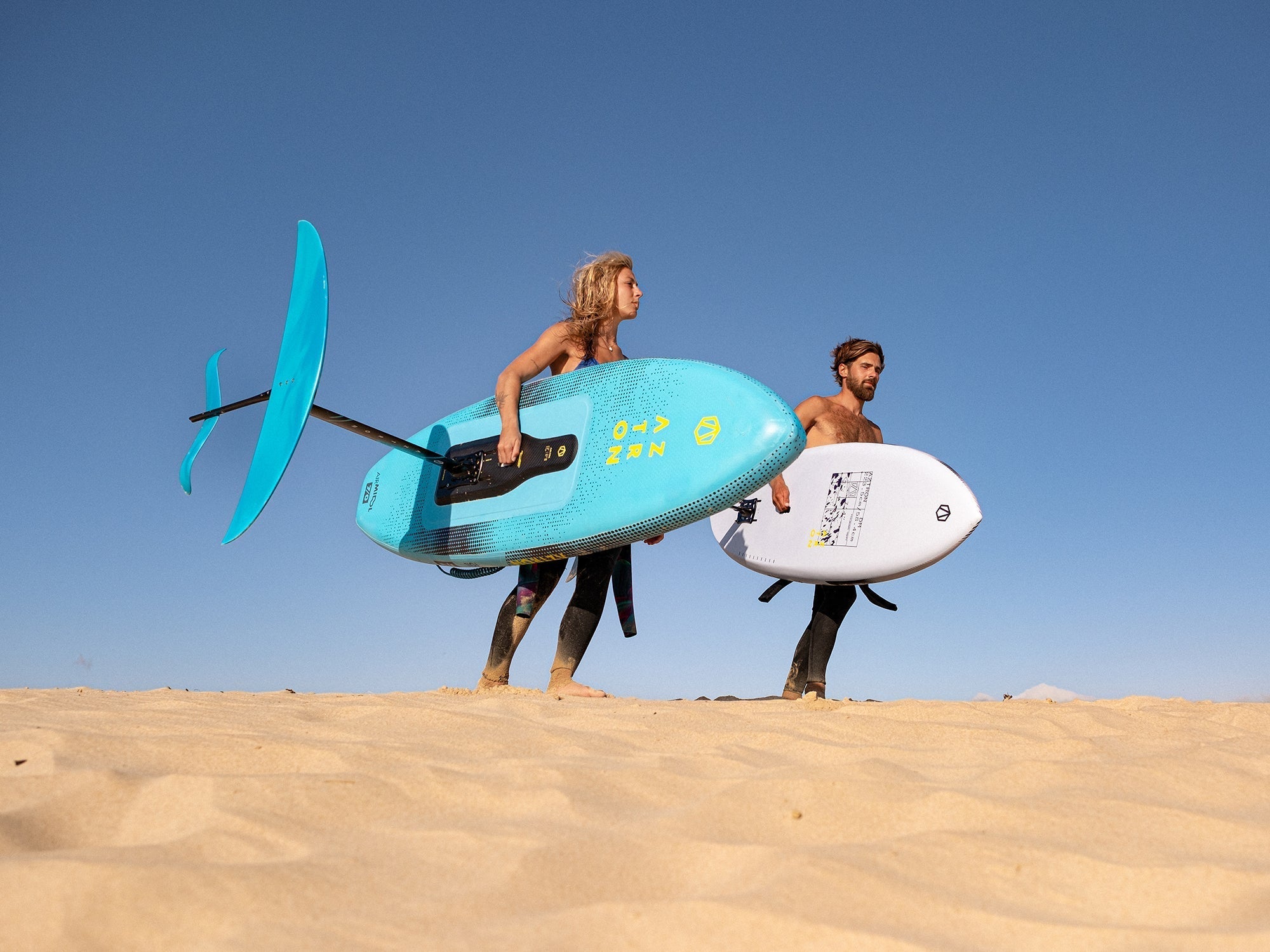Plajda mavi ve beyaz SUP board taşıyan çift, açık mavi gökyüzü altında