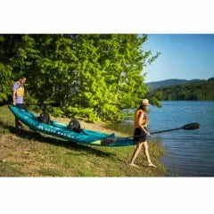 İki kişi, mavi Aqua Marina şişme kano ile göl kenarında açık hava aktivitesi yapıyor.