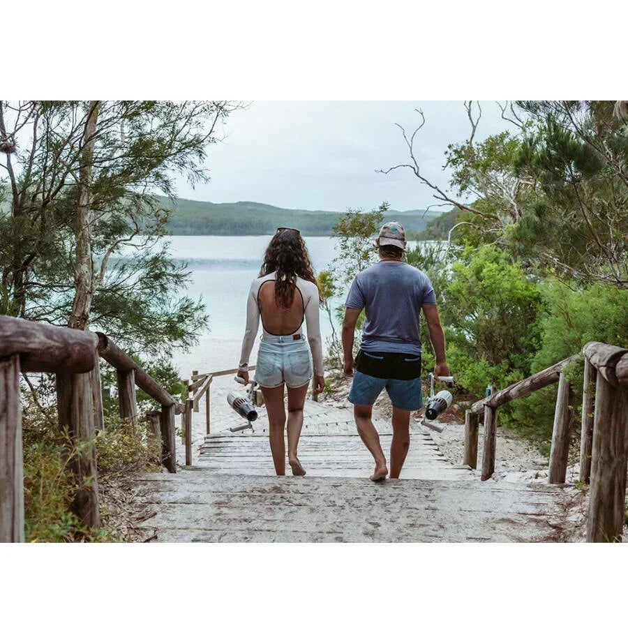 İki kişi, ellerinde su altı scooterı ile ağaçlıklı bir yolda göle doğru yürüyor.