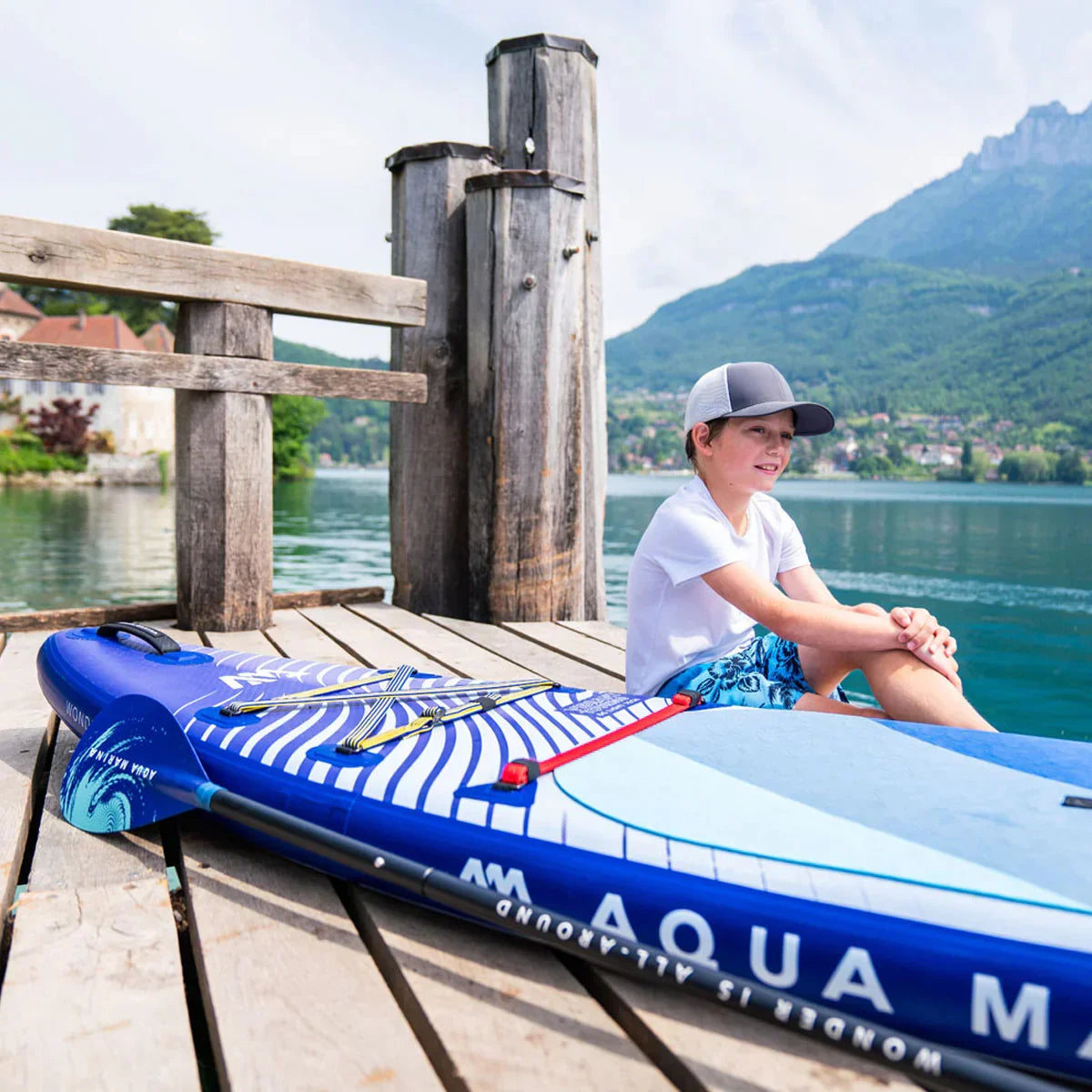 Gölde iskelede oturan çocuk, yanındaki mavi şişme SUP board ve doğa manzarası