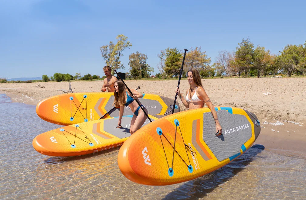 Plajda üç kişi, şişme turuncu SUP paddle board üzerinde kürekle suya giriyor.