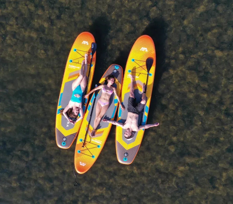 Üç kişi turuncu su üstü paddle board üzerinde gölde güneşleniyor ve dinleniyor.