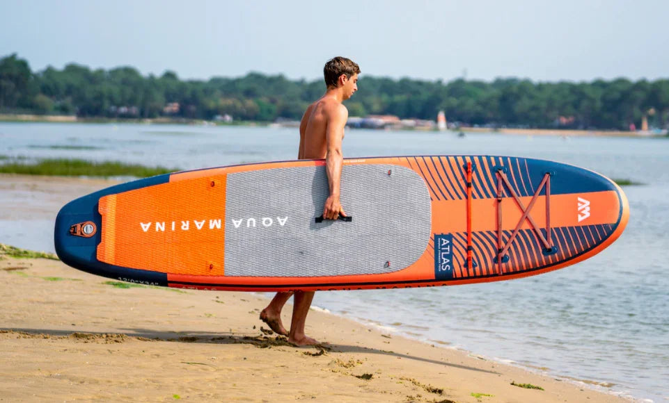 Plajda turuncu-gri Aqua Marina Atlas SUP board taşıyan bir adam, su sporları sahnesi
