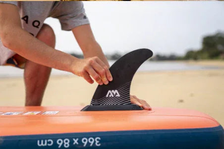 Portakal ve mavi renkli Aqua Marina Atlas şişme SUP board üzerine yüzgeç takılıyor, plaj fonunda.