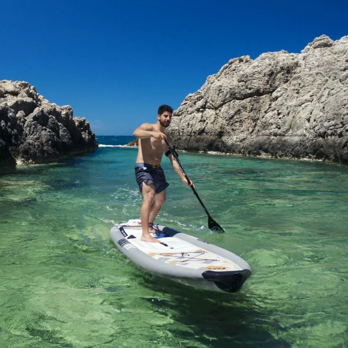 Turkuaz denizde, kaya manzaralı koyda Aqua Marina Drift SUP board üzerinde kürek çeken adam.