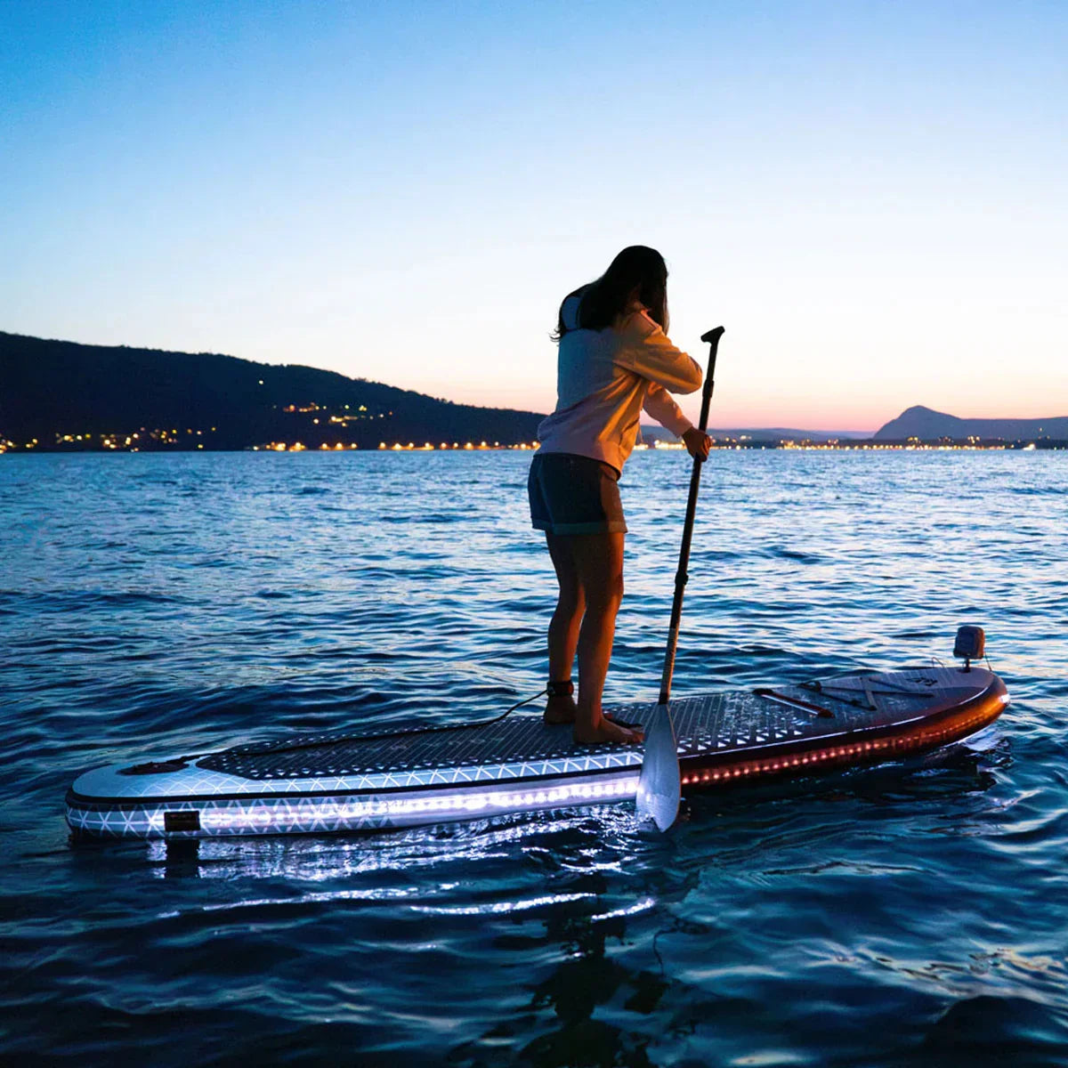 Gün batımında LED ışıklı SUP board üzerinde kürek çeken kişi, açık deniz ve manzara.