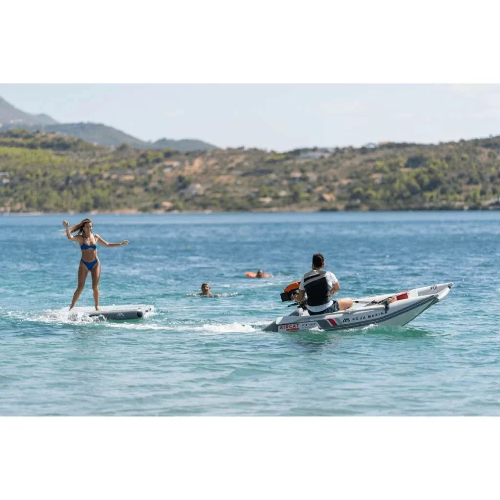 Denizde Aqua Marina Island Plus şişme platform üzerinde duran kadın, yanında Aircat motorlu bot ve yüzen kişiler, su sporu ve açık hava keyfi.