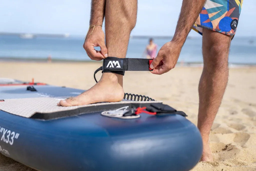 Kumsalda paddle board üzerinde ayağına güvenlik kayışı takan erkek, SUP board hazırlığı