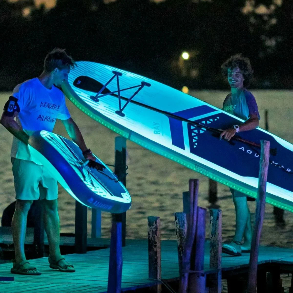 Gece isıklandırmalı Aqua Marina Ray SUP board ile iskelede iki kişi, arka planda su manzarası
