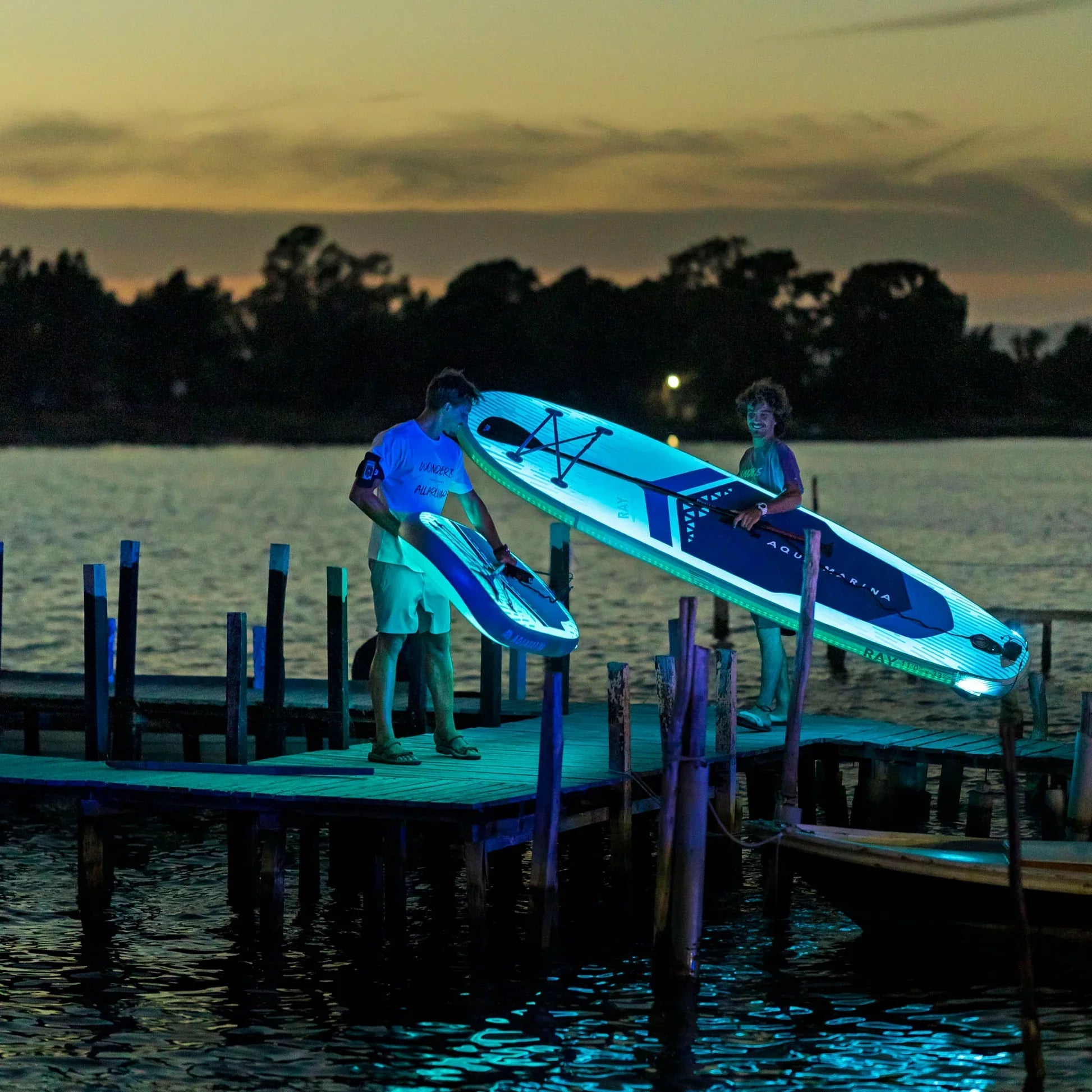Gün batımında iskelede Aqua Marina Ray SUP board taşıyan iki kişi, su sporları keyfi.