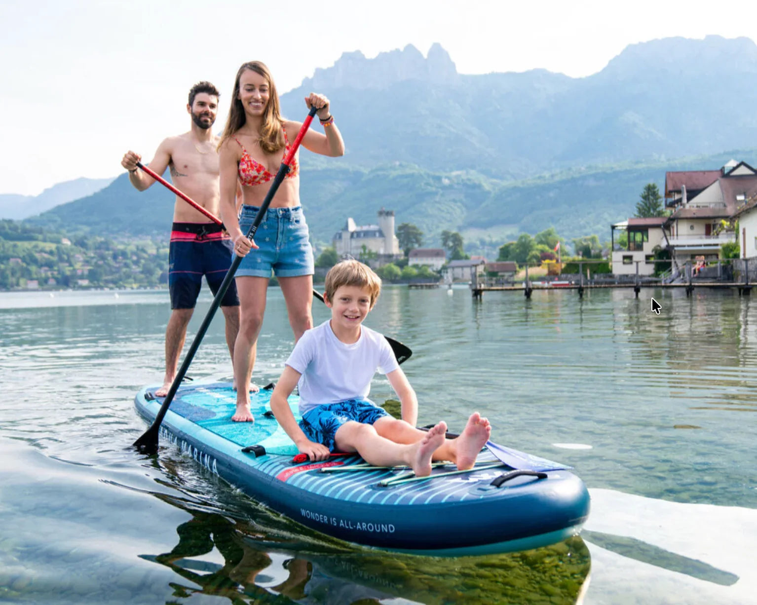 Gölde Aqua Marina SUP board üzerinde kürek çeken iki yetişkin ve oturan bir çocuk, arka planda dağlar ve evler.