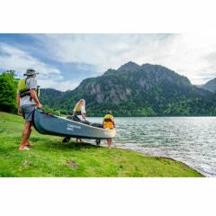 Üç kişi göl kenarında kano taşırken, arka planda dağ ve orman manzarası görülüyor.